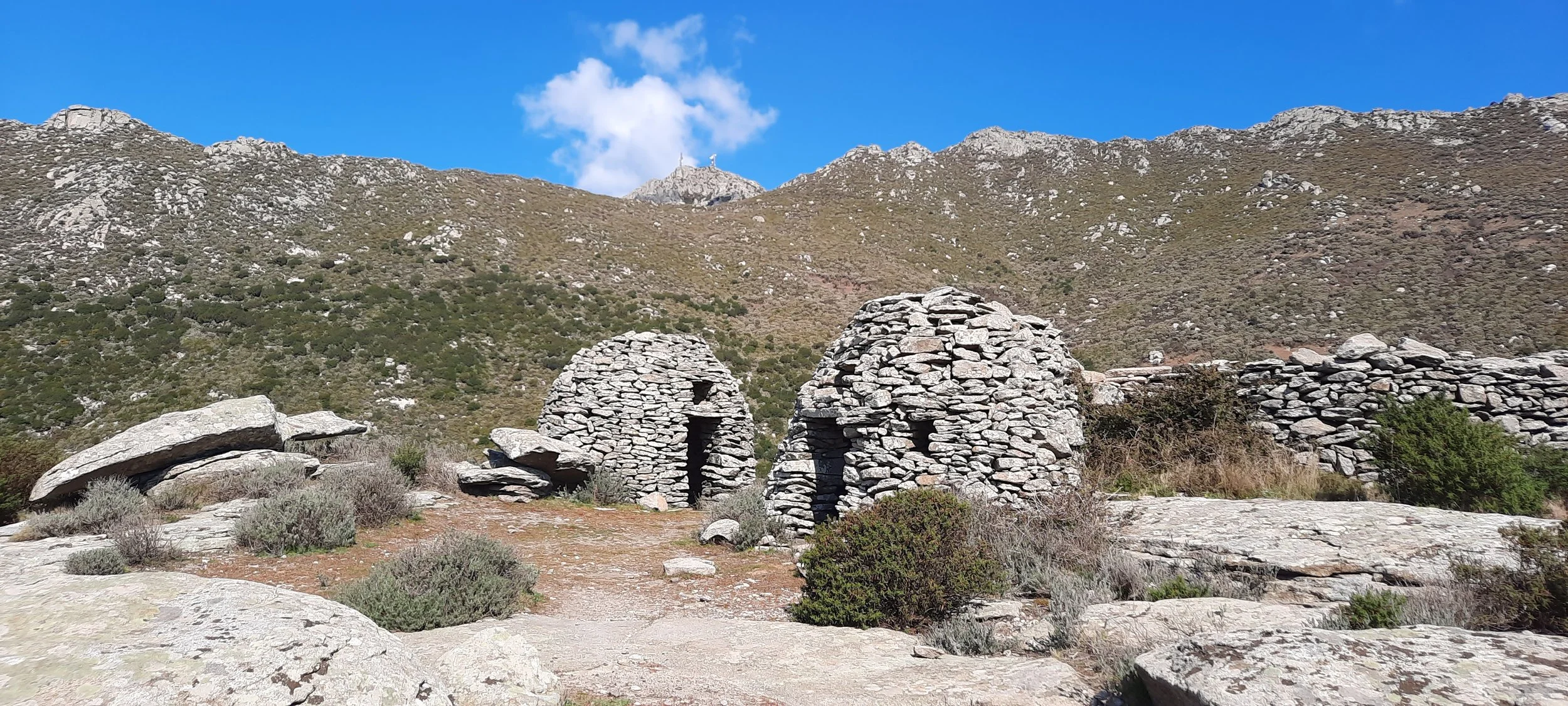 Due casette in pietra circondate da vegetazione di montagna con una catena montuosa sullo sfondo e un cielo blu con alcune nuvole.