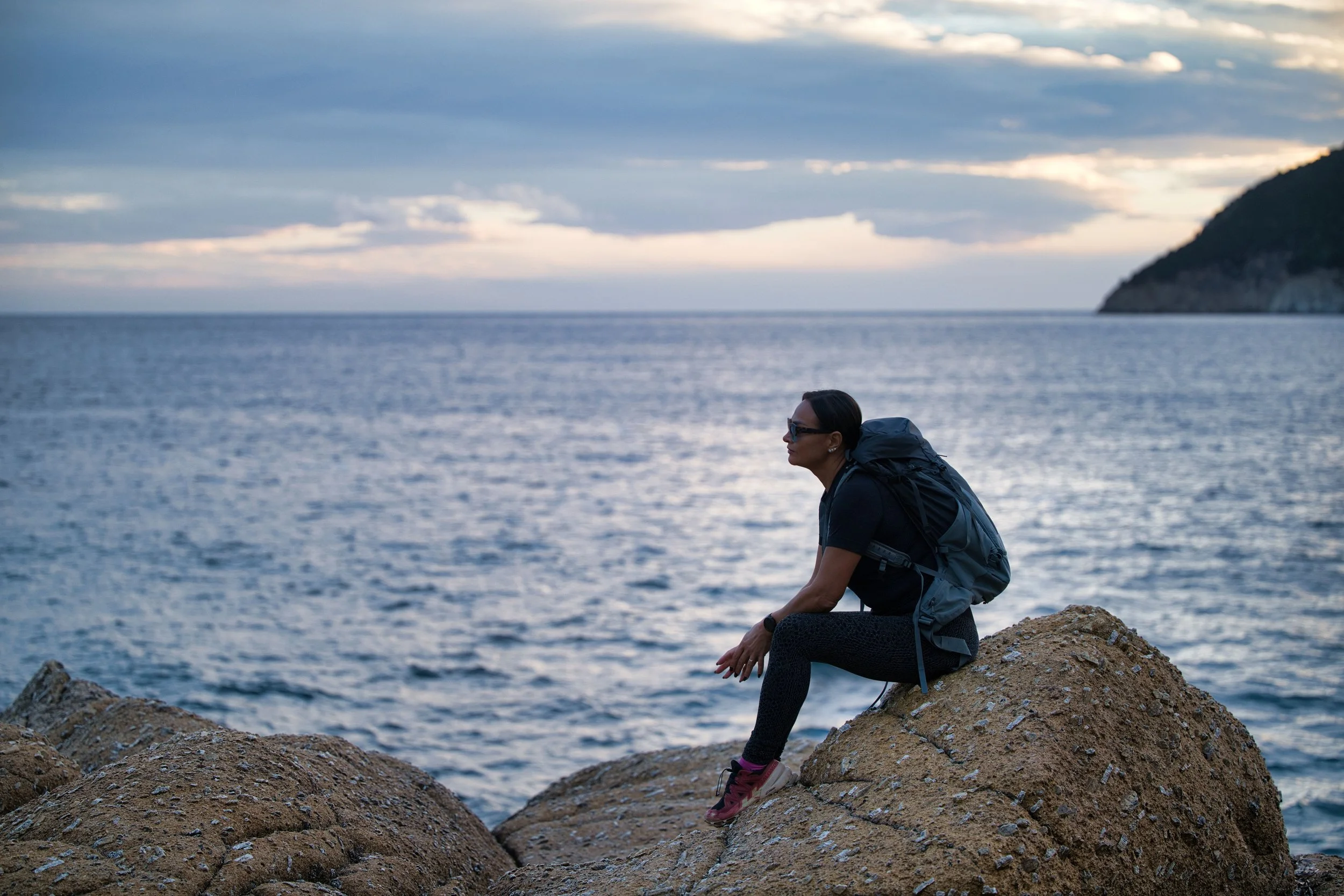 Donna seduta su una roccia vicino al mare al tramonto, indossa abbigliamento da escursionismo e ha uno zaino.