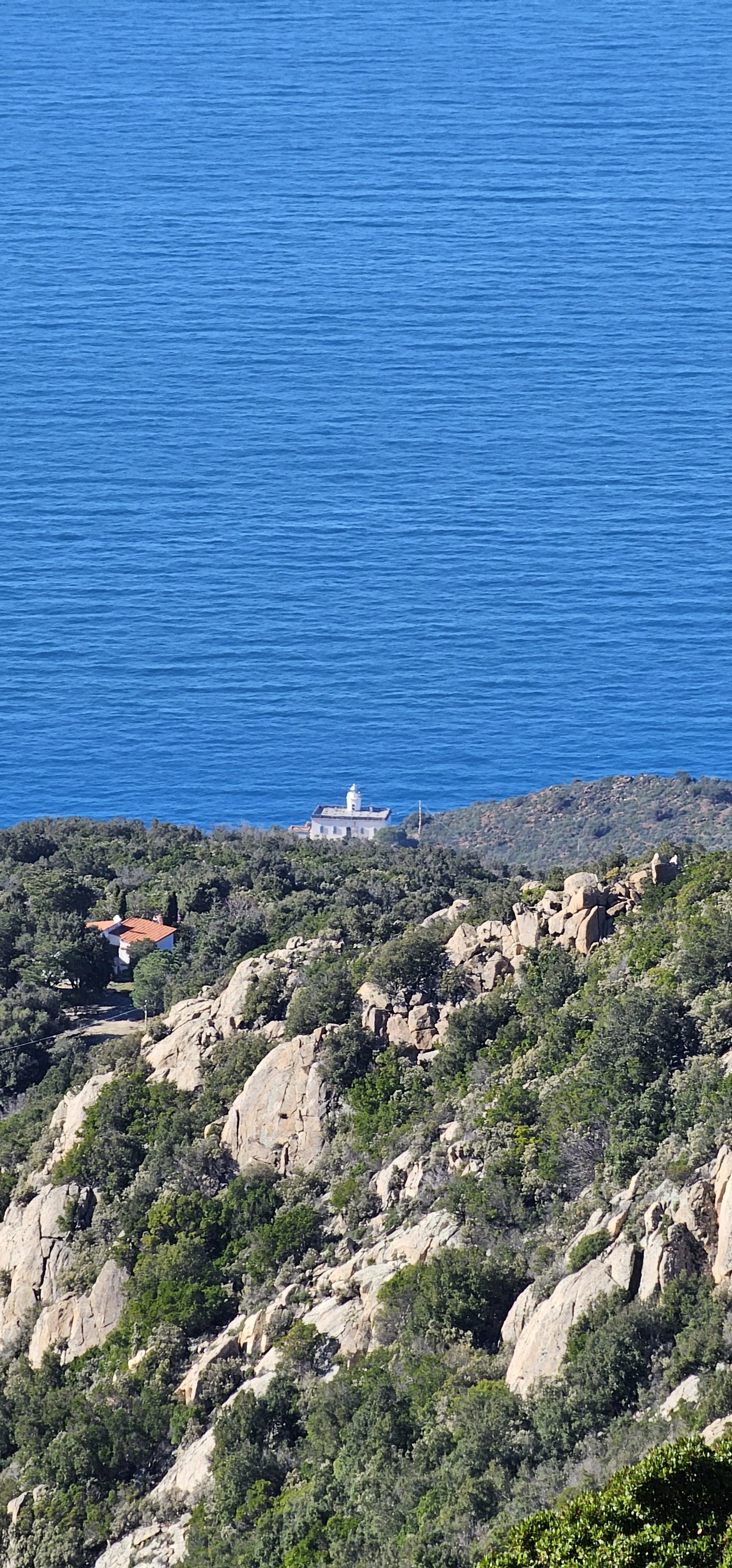 Paesaggio costiero con mare blu, scogliera rocciosa e vegetazione verde, e un faro bianco all'orizzonte