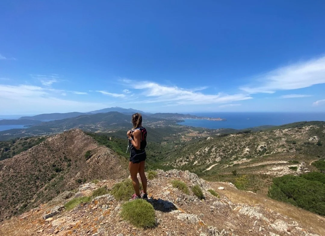Una donna in abbigliamento da trekking con uno zaino, che si trova su una cima, guarda un paesaggio montano e marino sotto un cielo blu con nuvole sparse.