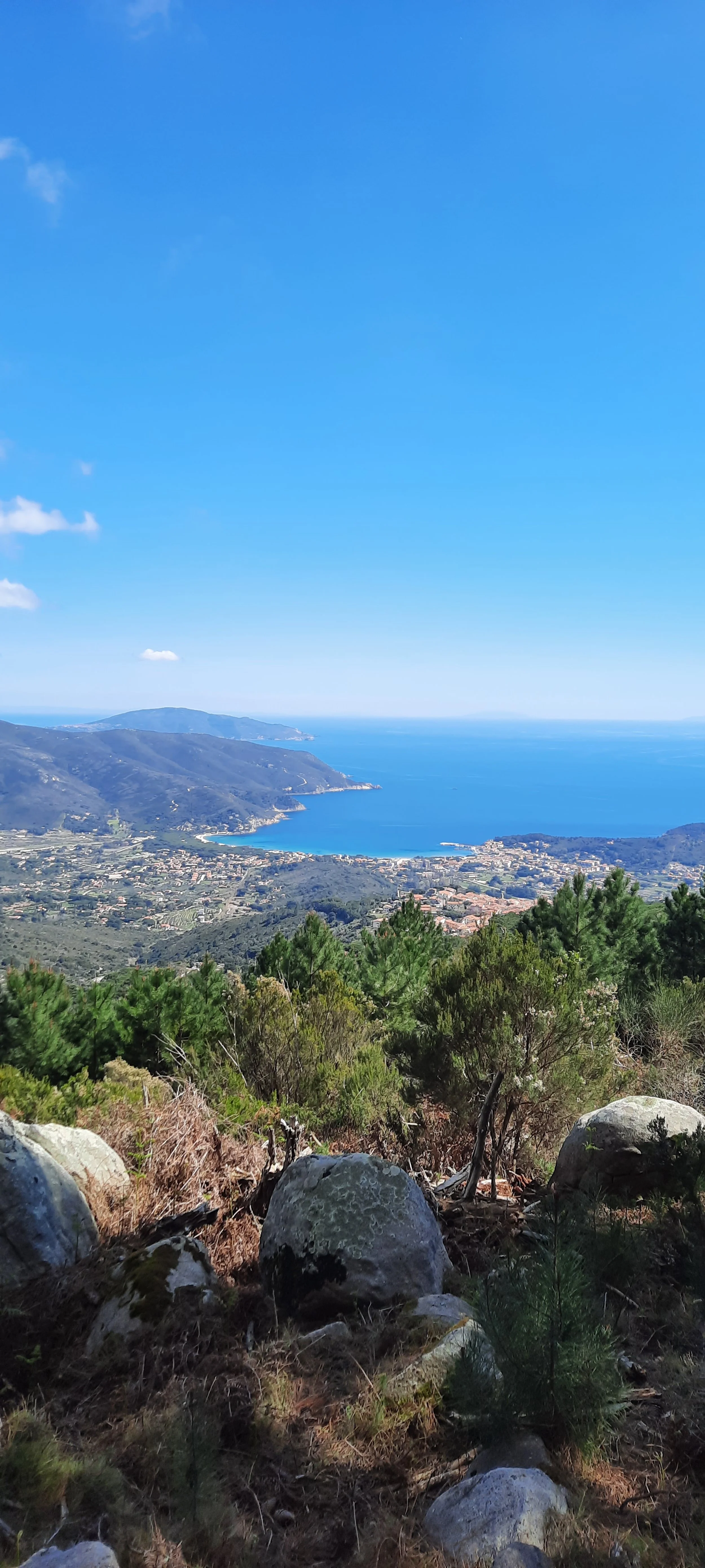 Vista di una costa con il mare blu, montagne verdi e un villaggio, vista dal alto tra alberi e rocce.