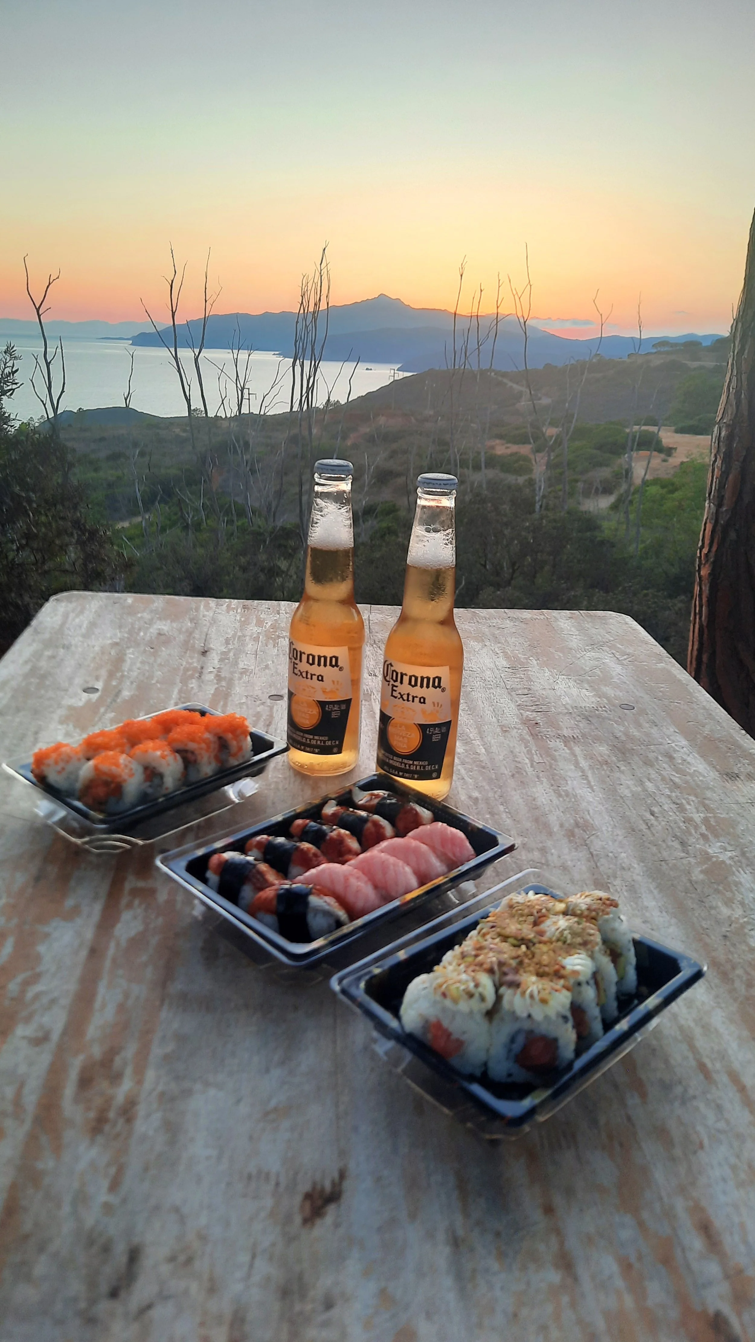 Sushi e due bottiglie di Corona Extra su un tavolo in legno, con vista al tramonto su monti e acqua, in un ambiente naturale.