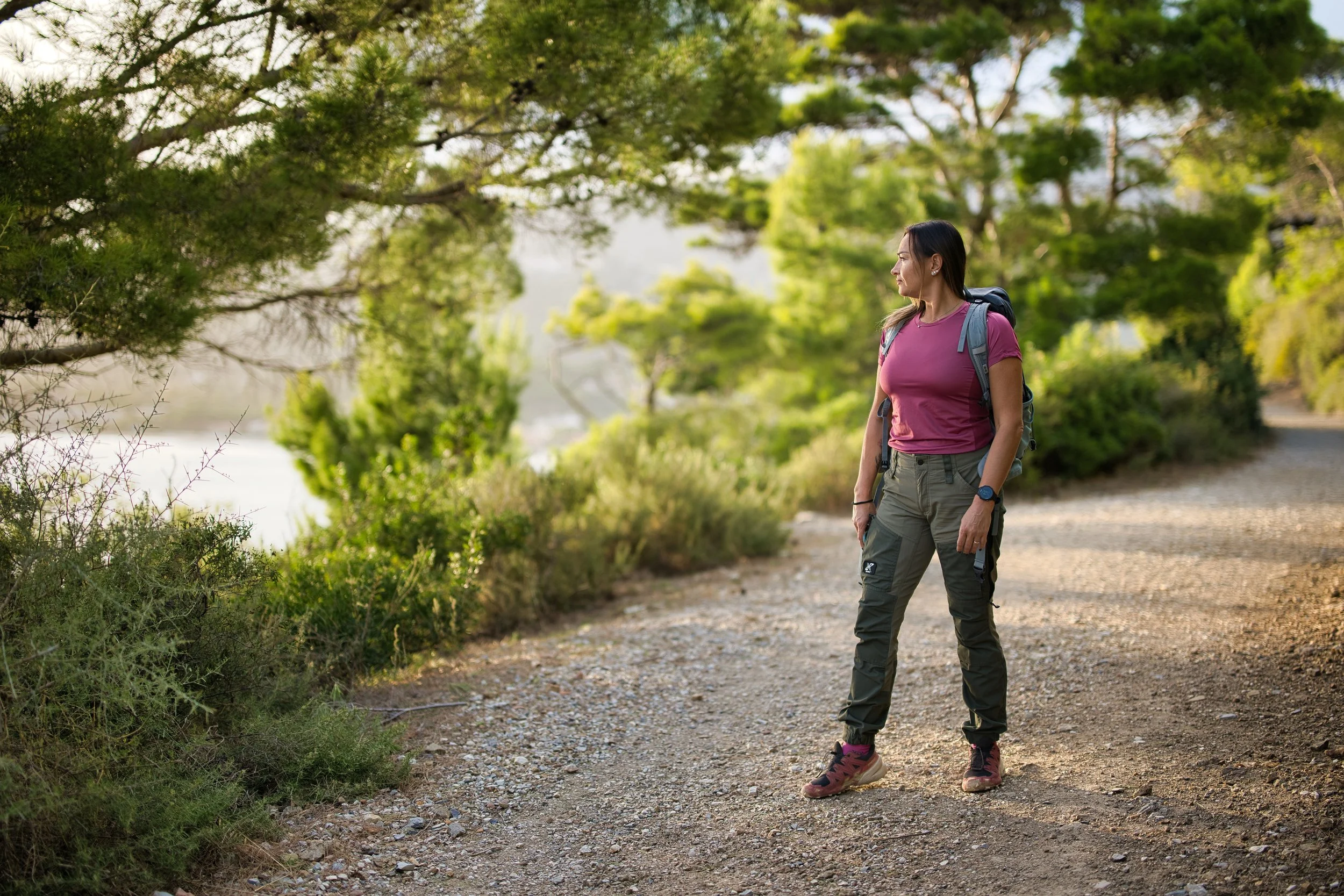 Donna con abbigliamento da escursionismo, zaino e orologio, in piedi su un sentiero immerso in una natura verdeggiante, con alberi e cespugli intorno, durante il giorno.