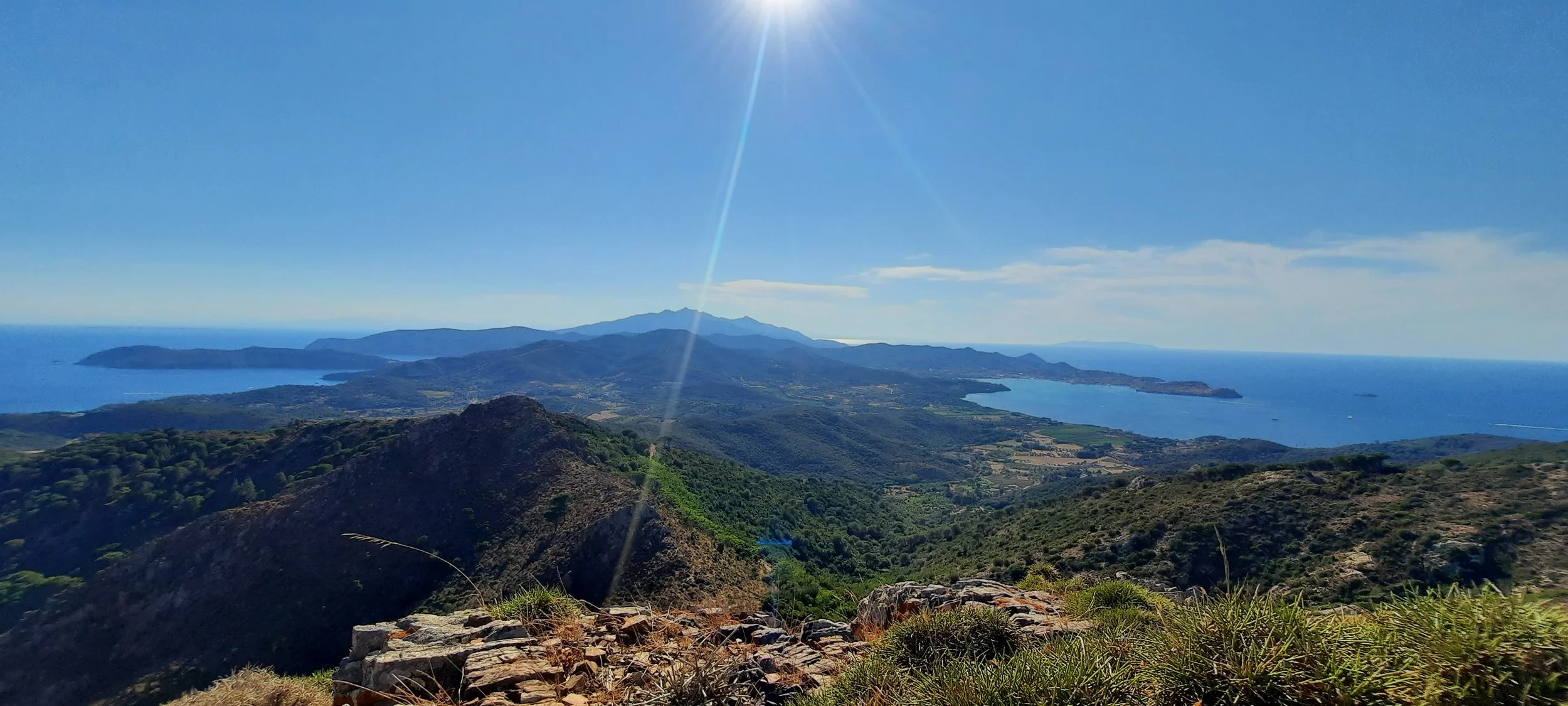 Panorama di colline e montagne con mare in lontananza sotto un cielo sereno e sole splendente