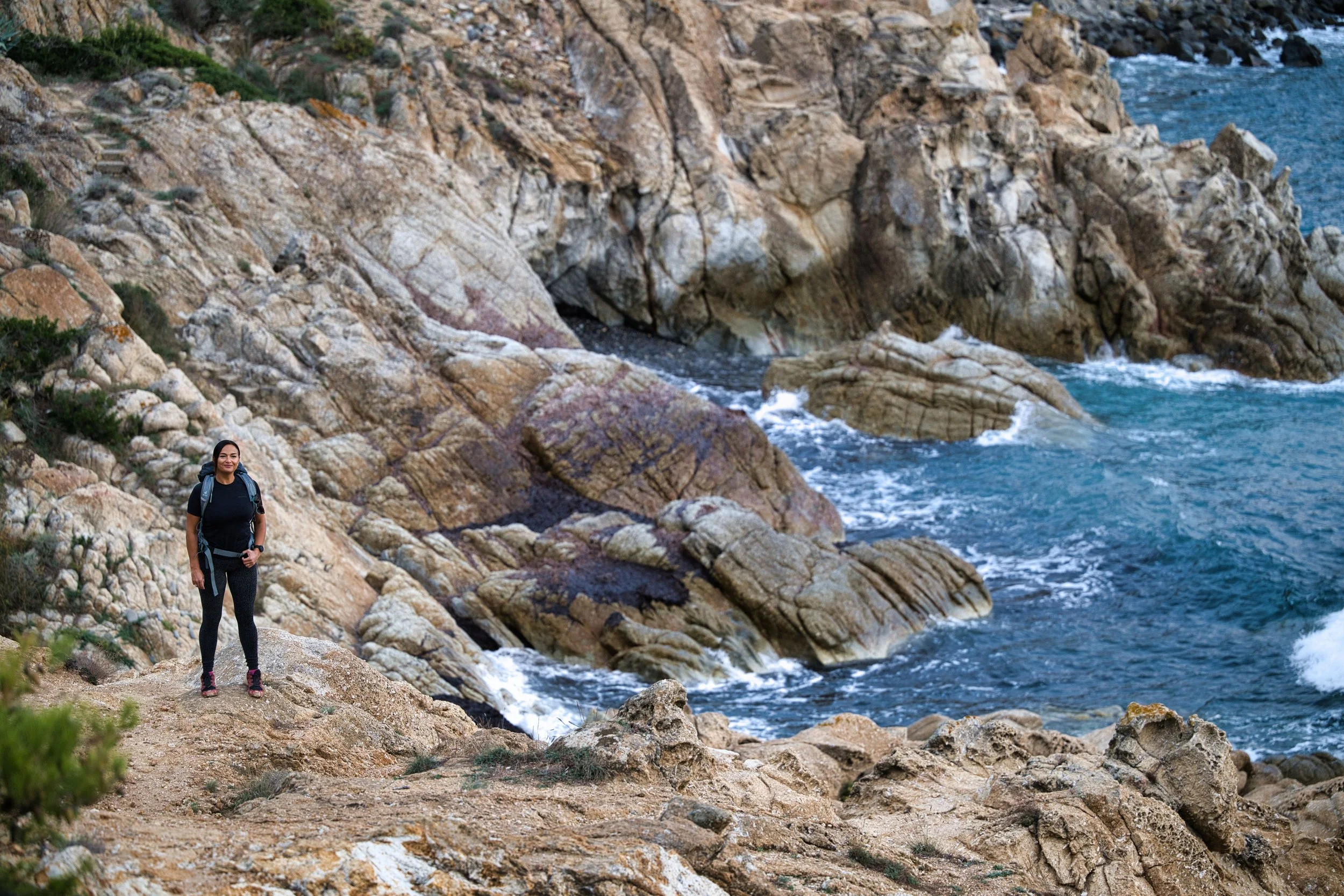Una donna con zaino cammina lungo un sentiero roccioso vicino al mare, con onde che si infrangono contro le rocce.