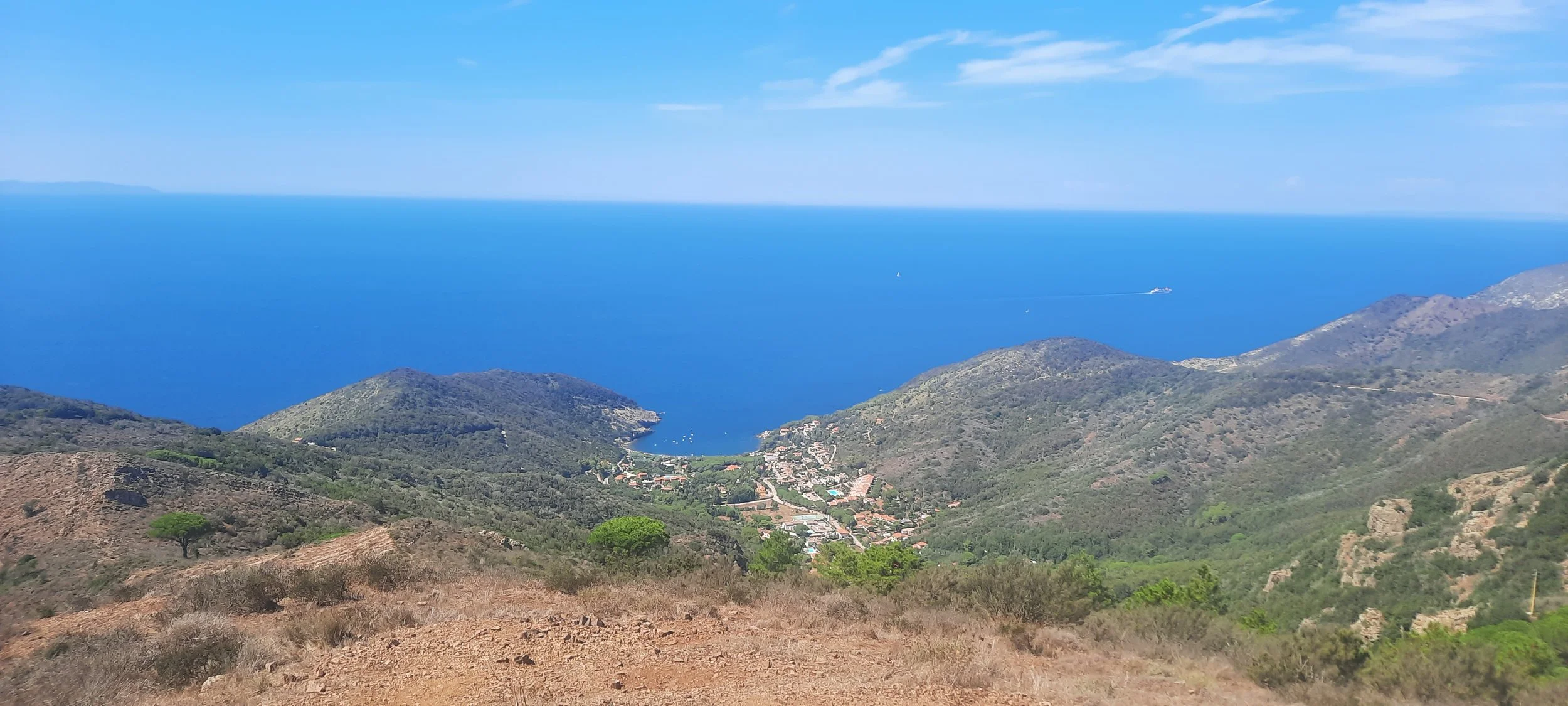 Vista panoramica di un mare blu con colline verdi e un piccolo villaggio vicino alla costa, sotto un cielo sereno.