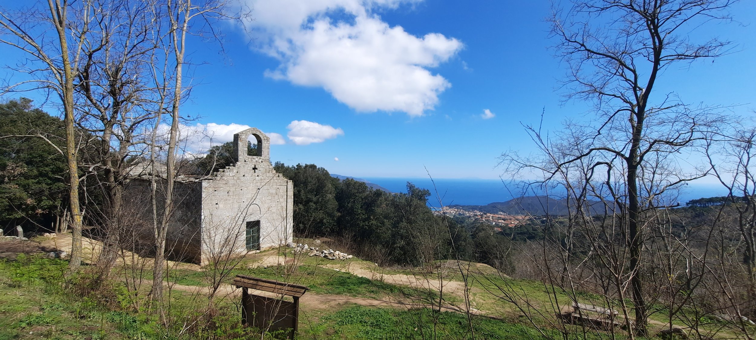 Piccola chiesa di pietra con campanile, circondata da alberi spogli, con vista sul mare e sulle colline sottostanti sotto un cielo blu con nuvole bianche.