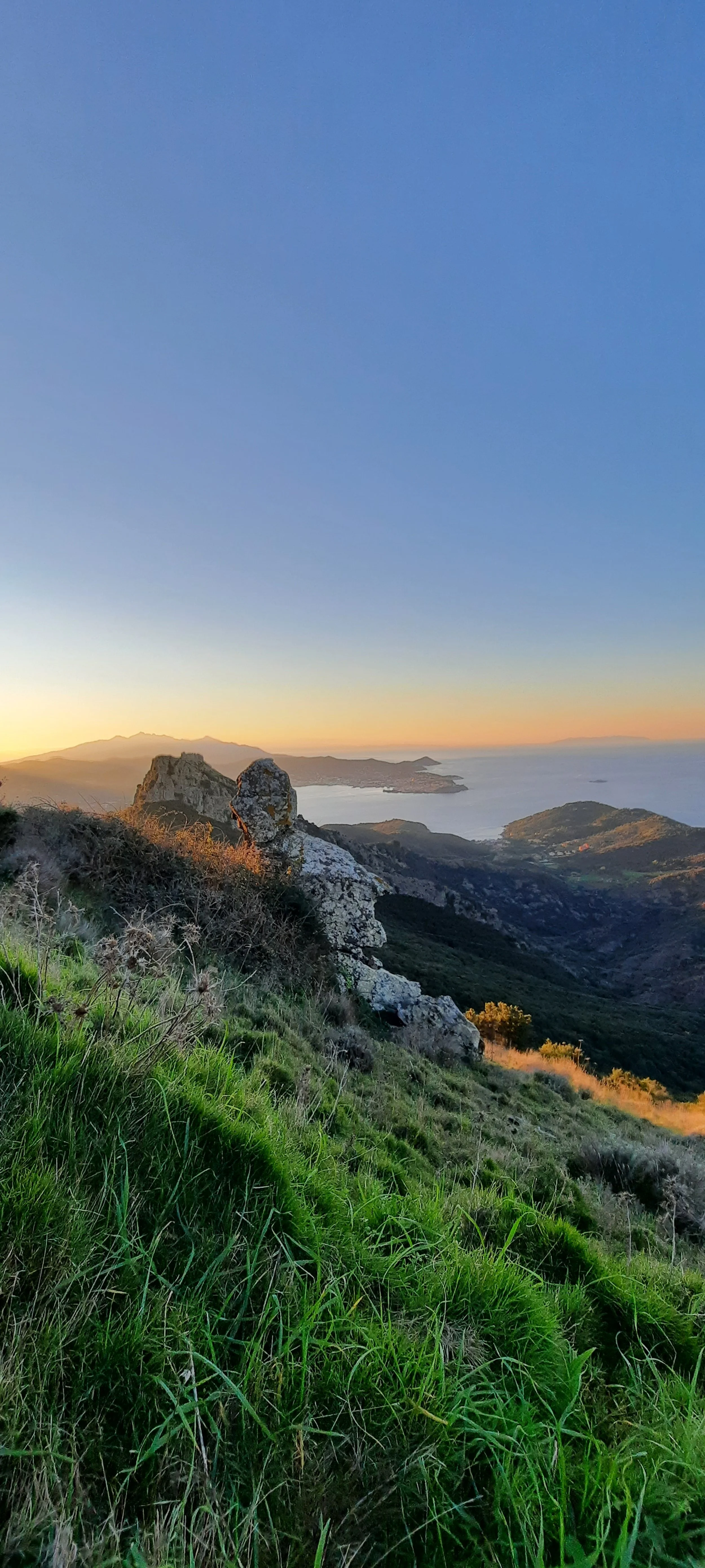 Paesaggio montano con rocce, vegetazione verde e vista sul mare durante il tramonto.