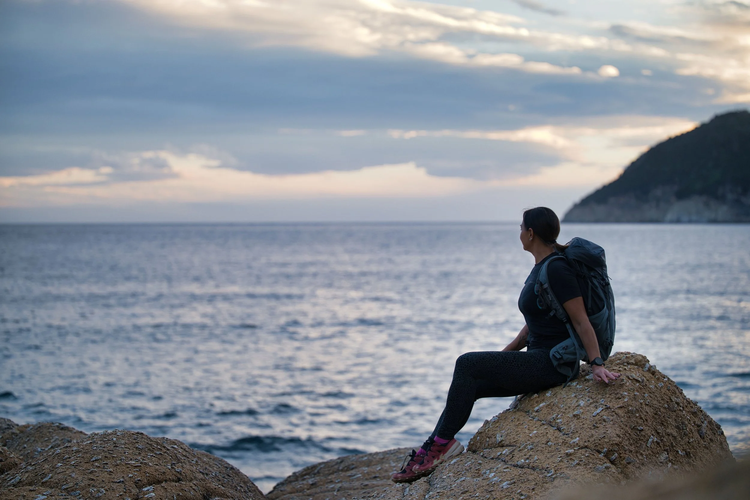 Una donna con uno zaino sportivo seduta su una roccia che guarda il mare al tramonto, con una collina sullo sfondo.