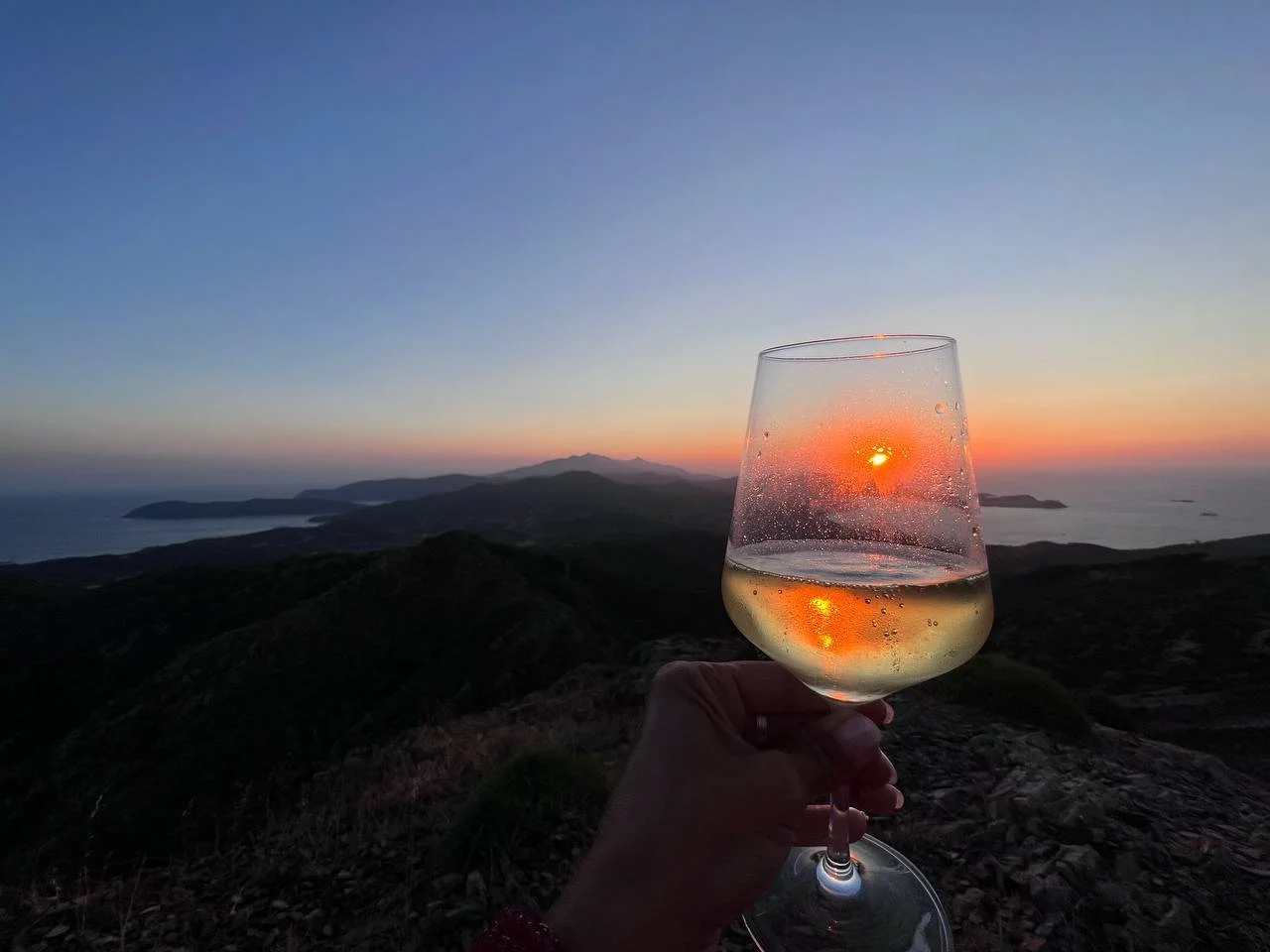Calice di vino bianco con vista su un tramonto sul mare e le montagne.