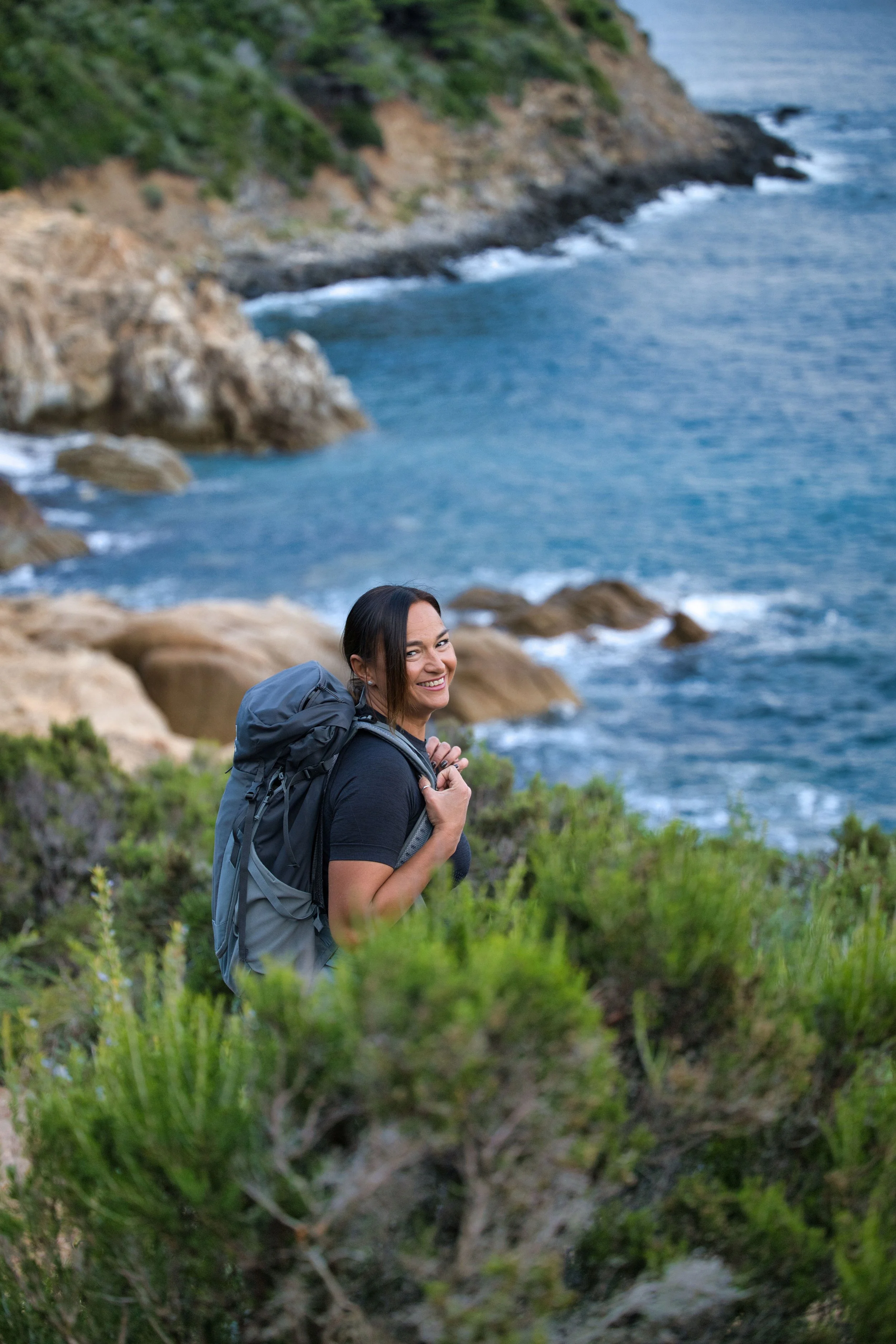 Una donna con uno zaino in piedi vicino al mare, circondata da vegetazione, con uno sfondo di scogliera e onde che si infrangono sulla riva.