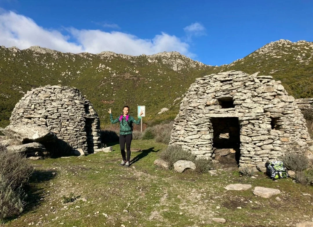 Due strutture in pietra costruite con pietre incastonate, un'alpinista che fa un gesto di vittoria, uno sfondo di montagne verdi e cieli azzurri.