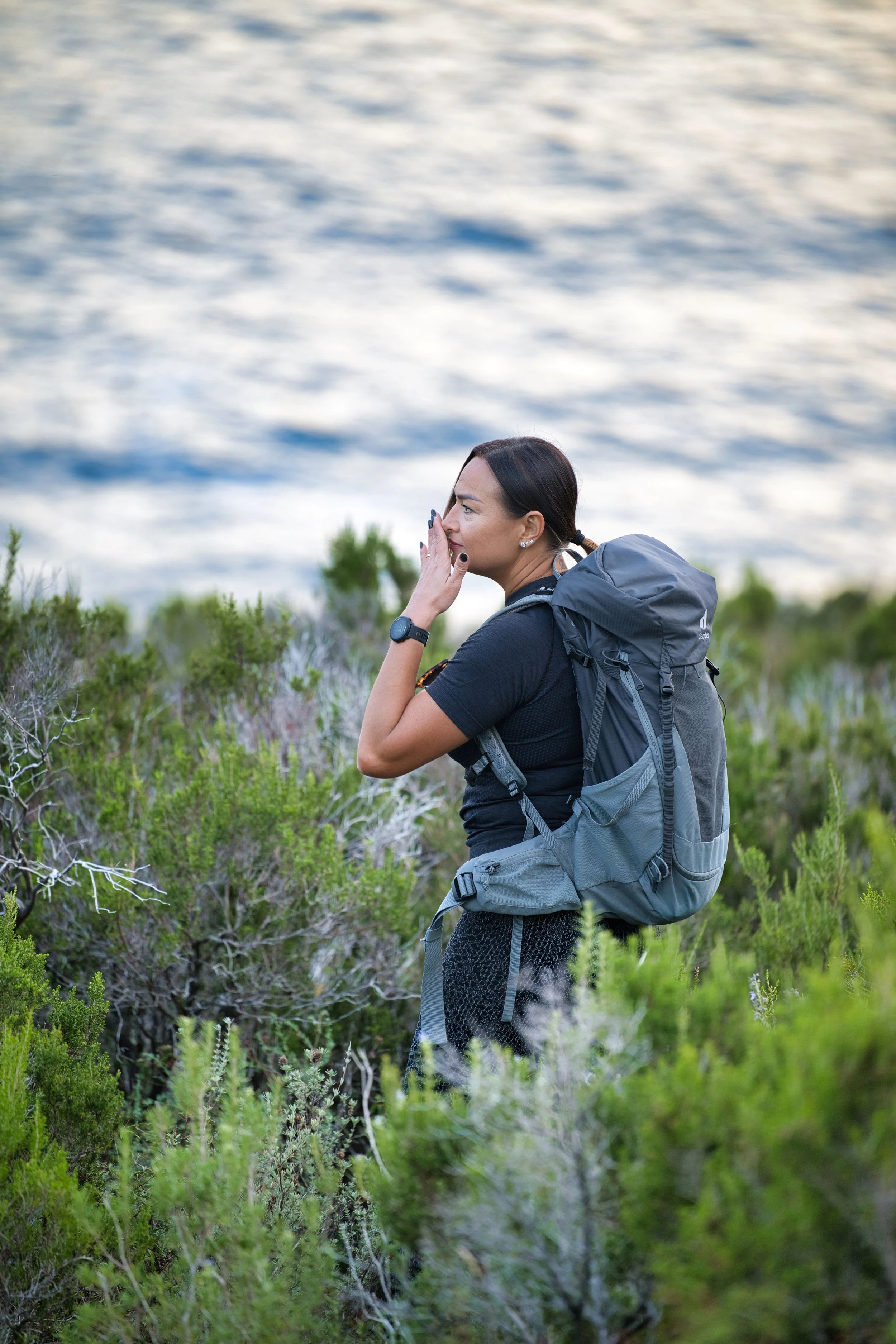 Donna con zaino grande che cammina tra arbusti vicino a un lago o mare