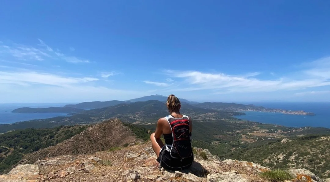 Una donna con uno zaino seduta su una roccia in cima a una montagna, con vista panoramica su coste, mare e colline sotto un cielo azzurro.