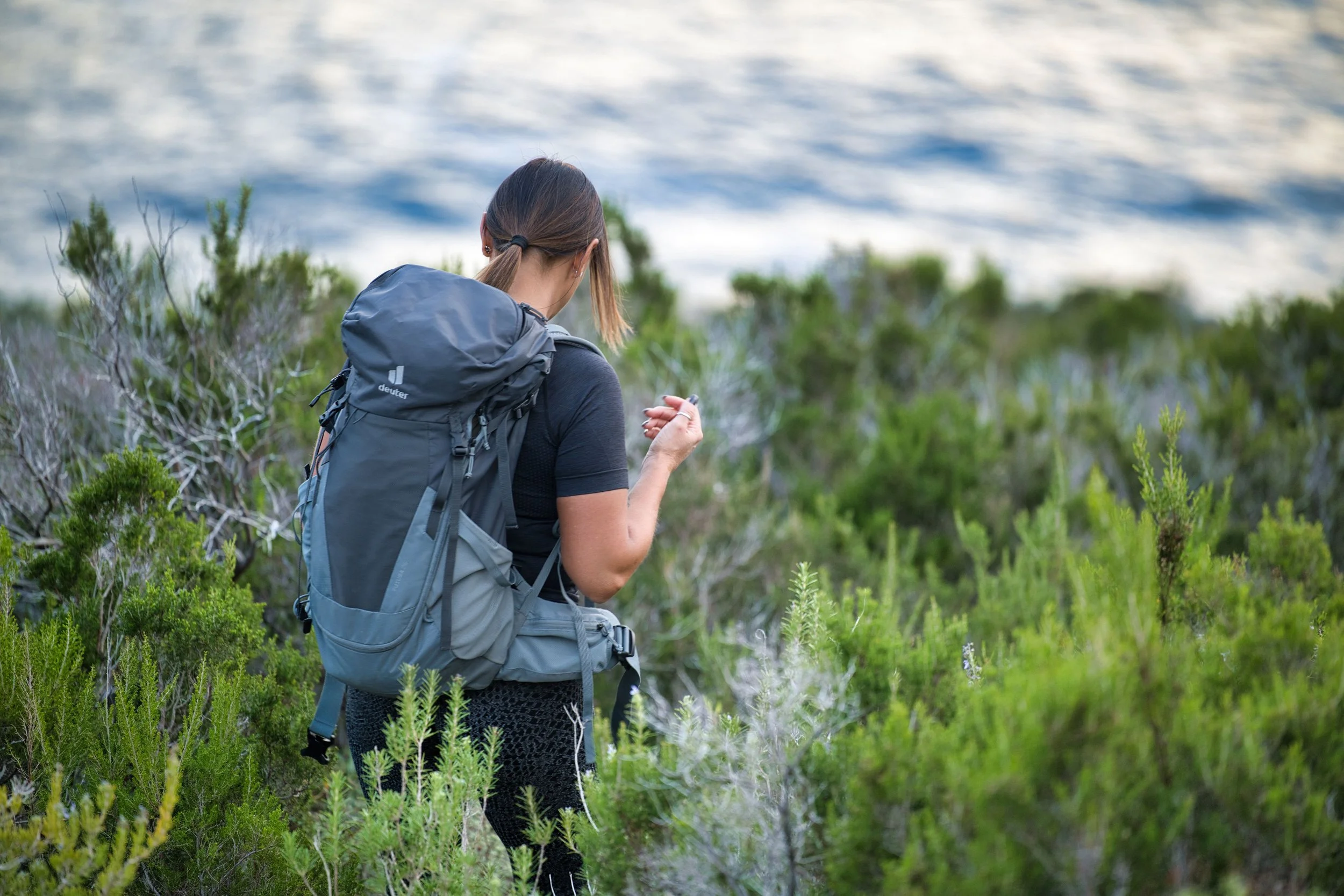 Una donna con uno zaino grande sta camminando tra la vegetazione vicino al mare.