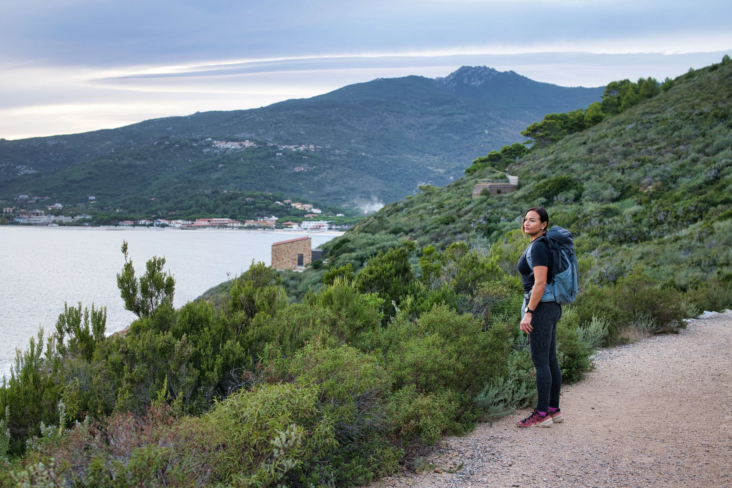 Una donna con zaino che cammina lungo un sentiero in un paesaggio naturale con colline e acqua sullo sfondo