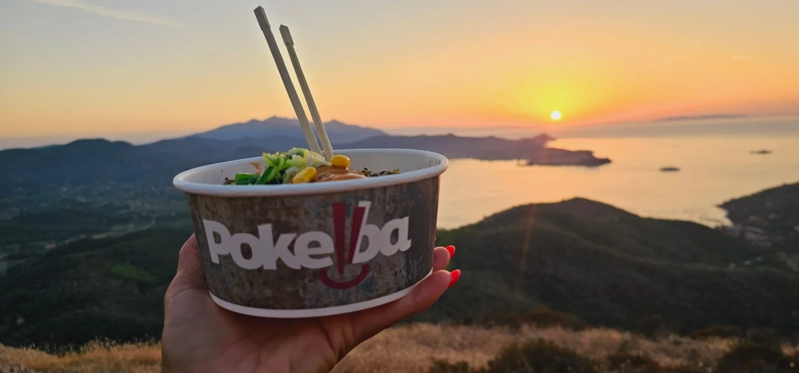 Una mano che tiene una ciotola di cibo con lo sfondo di un tramonto sopra il mare e le isole, con un paesaggio montano sottostante.