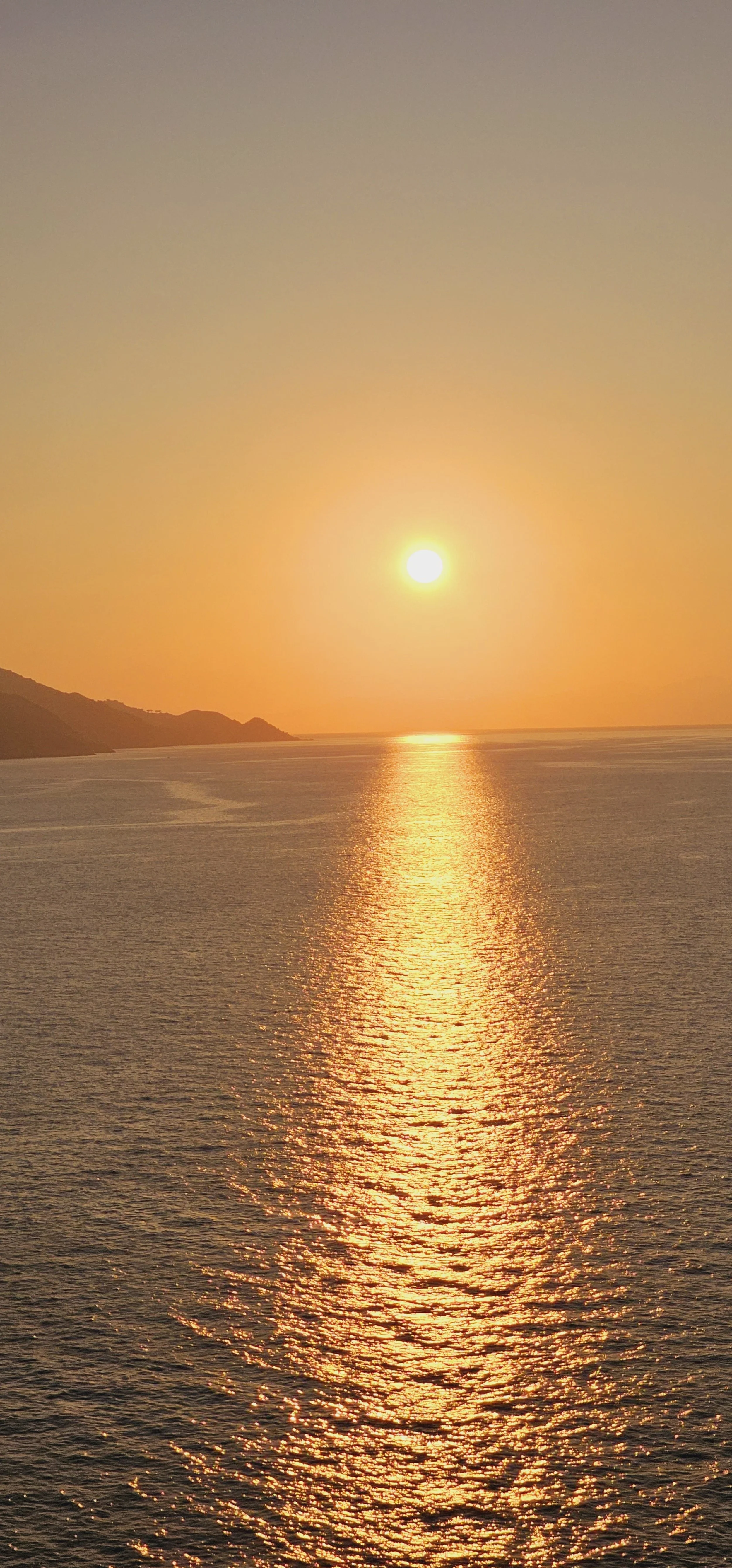 Tramonto sul mare con riflesso dorato sull'acqua e silhouette di colline sullo sfondo.