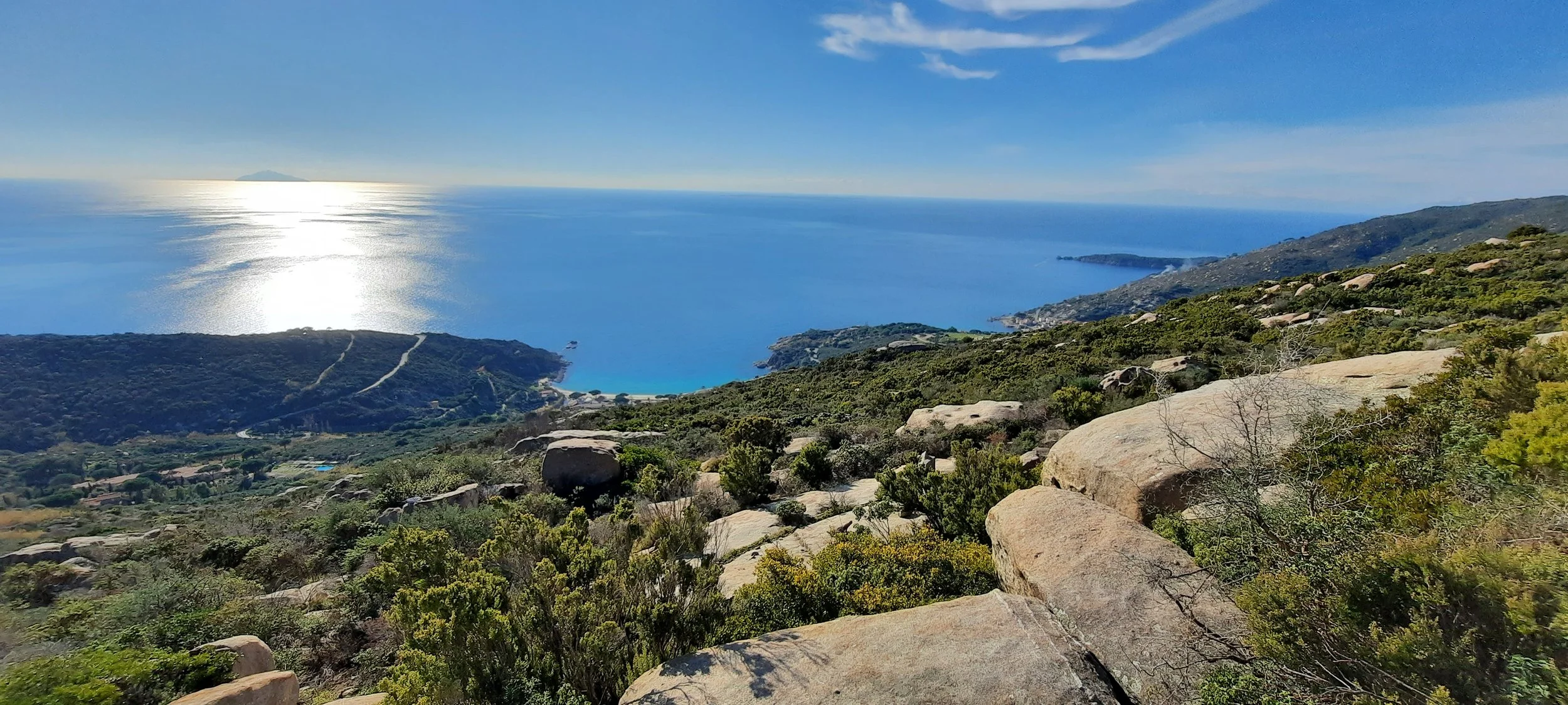 Paesaggio costiero con mare blu, colline verdi, rocce e vegetazione con cielo soleggiato.