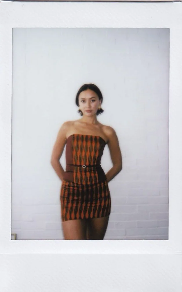 A woman posing against a white brick wall, wearing a strapless orange and brown patterned dress with a black belt.