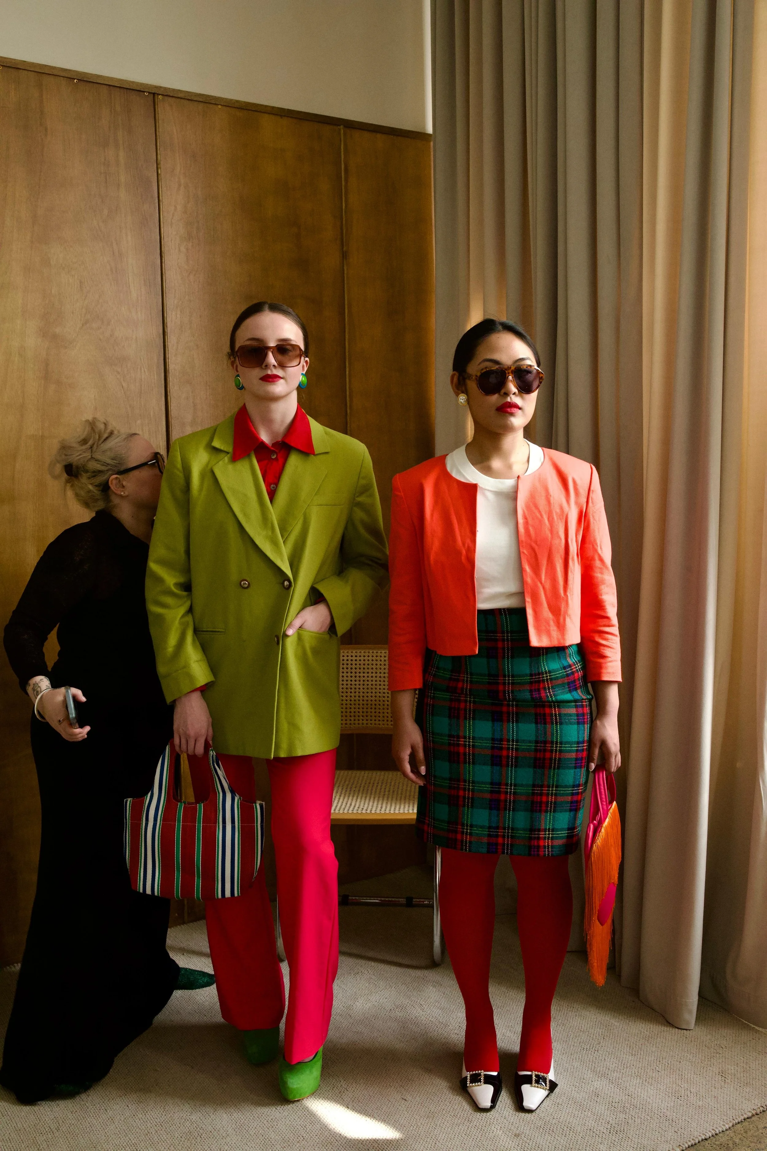 Two women standing indoors in front of a curtain, wearing colorful outfits and sunglasses. One is dressed in a green blazer with red pants, and the other in a red jacket with a plaid skirt. A stylist wearing black fixes the back of the green blazer