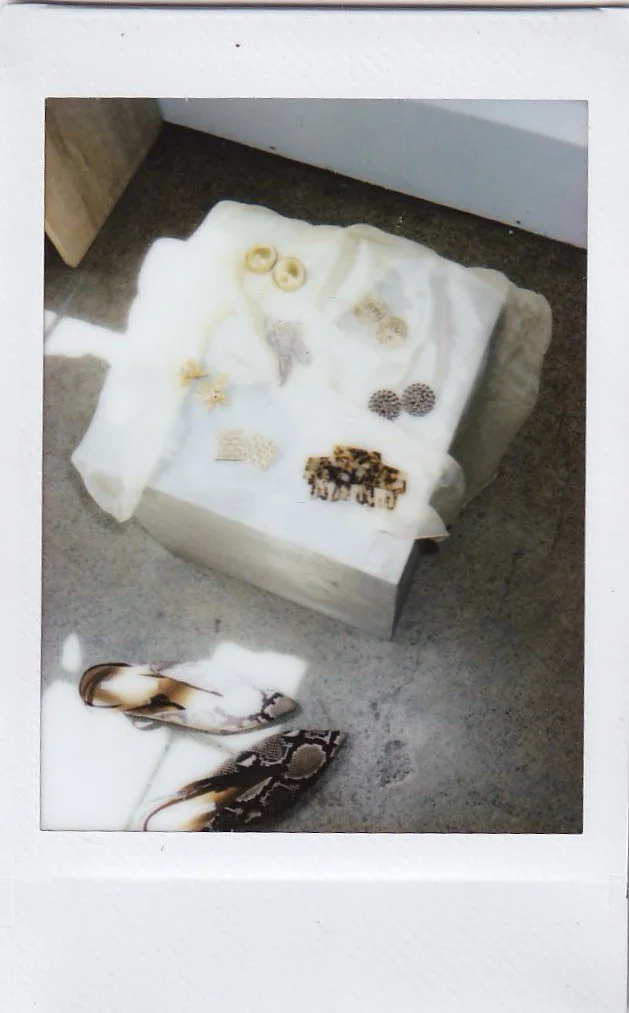 A white crate or box with various jewelry, including earrings and necklaces, on a white cloth. A pair of snakeskin-patterned shoes on the floor nearby.