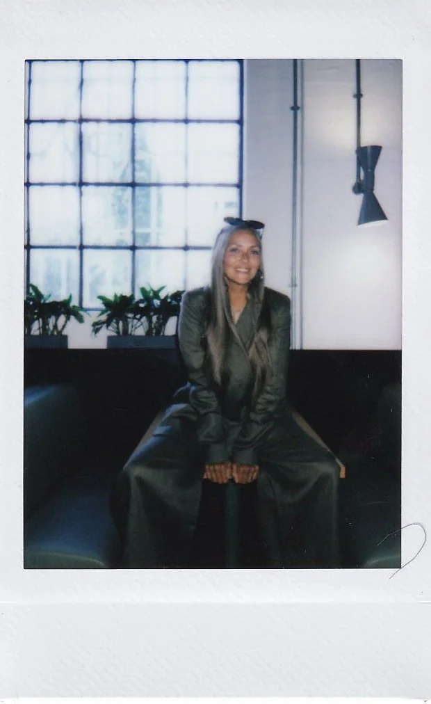 A woman with long gray hair sitting on a black bench in front of a large window with grid panes, smiling, wearing a dark green suit with cat ears on her head, and plants on the window sill behind her.