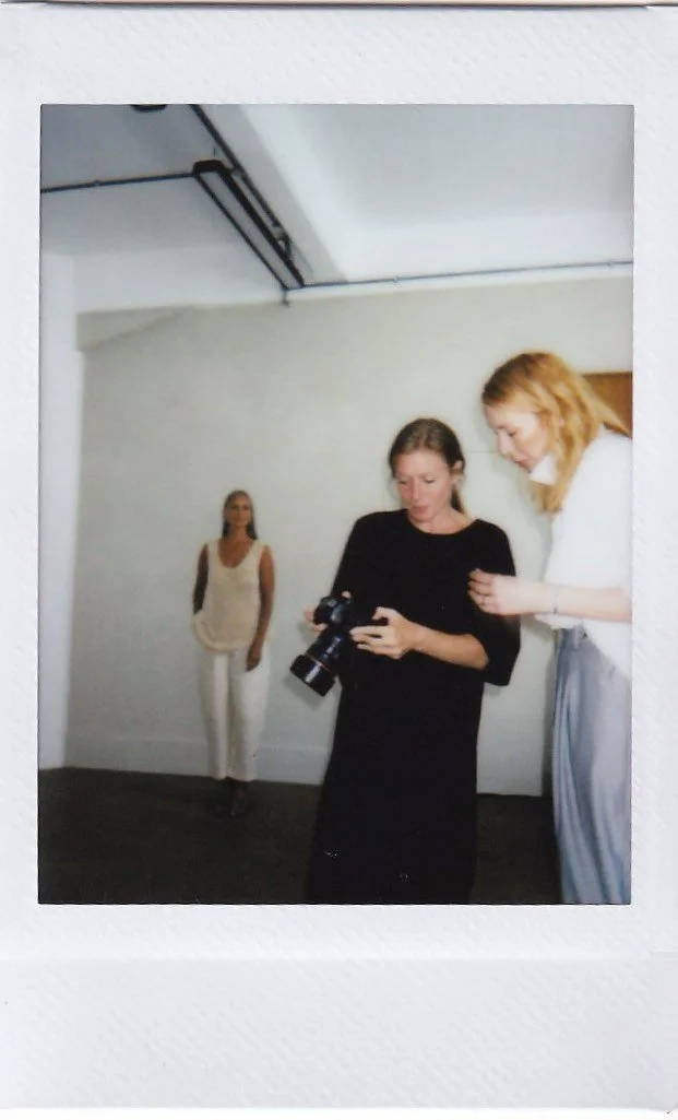 Three women in an art gallery or studio, one holding a camera, two engaged in conversation, with a plain white wall in the background.