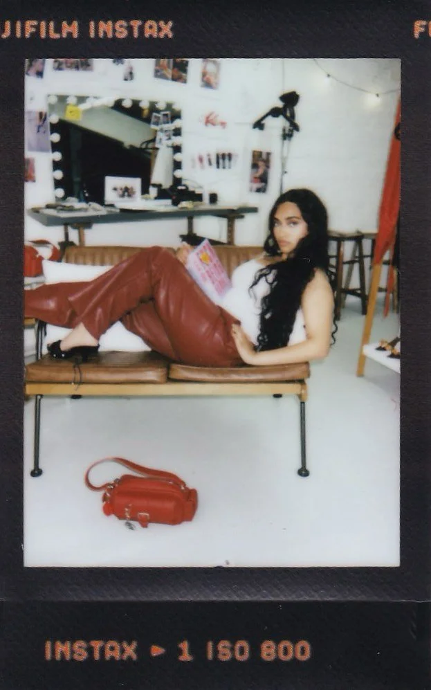 Young woman with long dark hair reclining on a bench in a stylish room, holding a magazine, with a red purse on the floor in front of her.