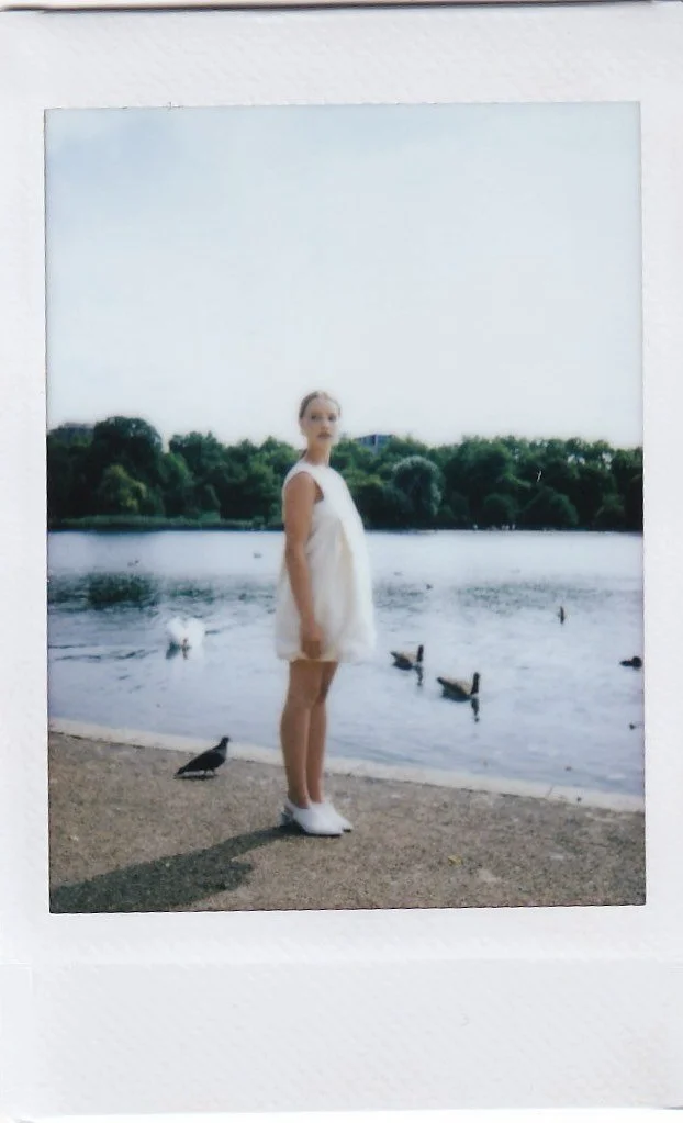 A young woman in a white dress standing by a lake with ducks, trees, and a cloudy sky in the background.