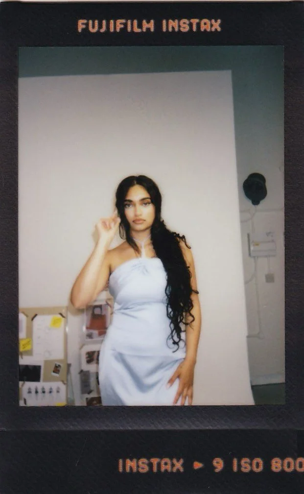 A woman with long, wavy black hair wearing a white strapless dress, standing indoors against a plain wall.