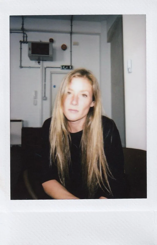 A woman with long blonde hair sitting indoors, wearing a black top, with a slightly tilted head and a subtle smile.