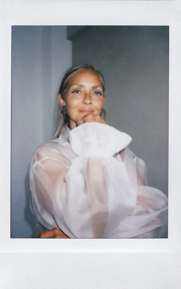 A woman with blonde hair and blue eyes wearing a cream-colored sheer blouse with puffed sleeves, posing indoors with her hand resting under her chin and smiling slightly.