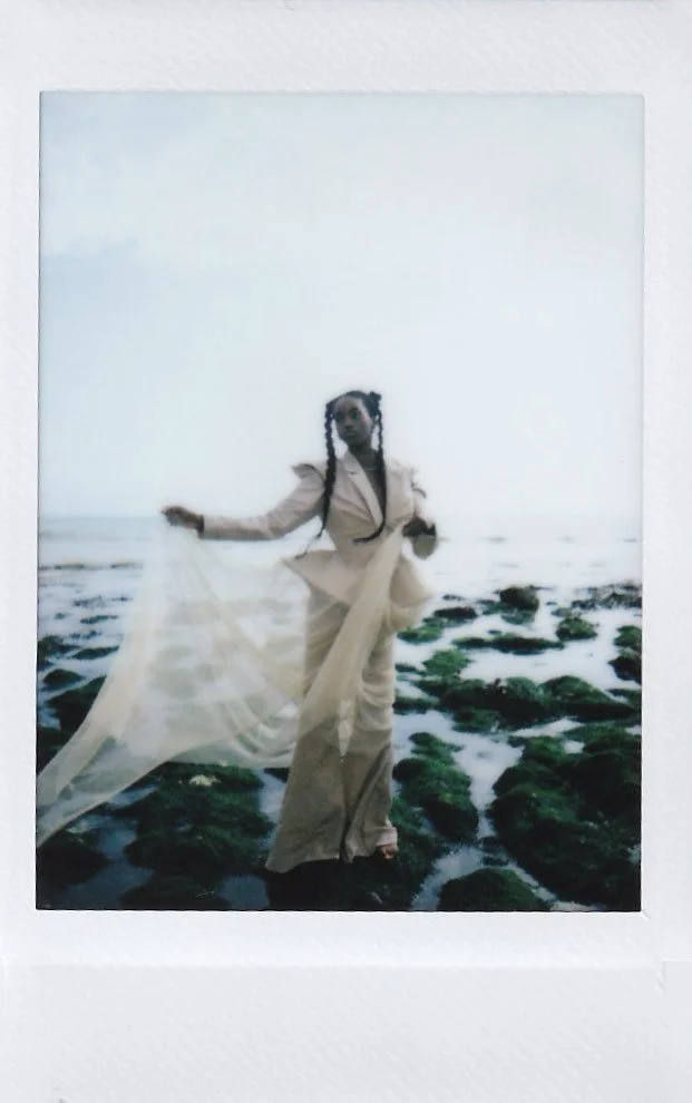 A woman stands on a rocky shoreline with the ocean in the background, wearing beige clothing and holding a flowing sheer fabric.