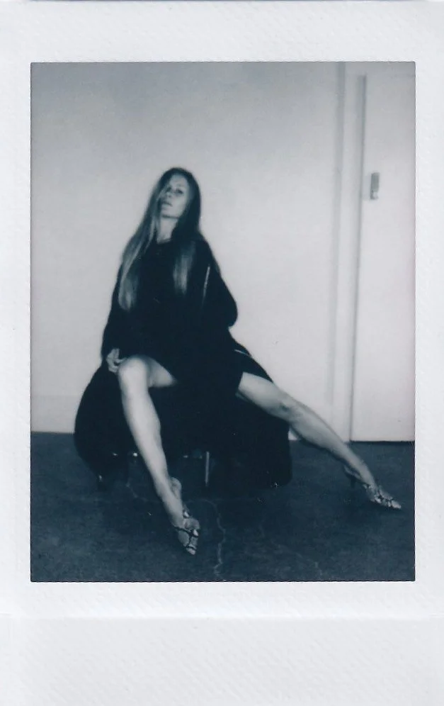 A woman sitting on a chair with her legs extended, wearing high heels and a black outfit, with long hair, in a room with a plain wall and a closed door.