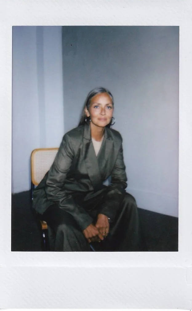 A woman with shoulder-length hair and earrings, dressed in a gray suit, sitting on a chair in a simple room with plain walls.