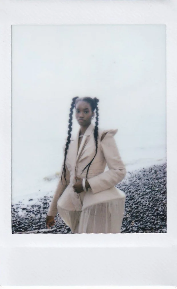 A woman with long, braided hair standing on a black pebble beach near the water, wearing a beige blazer and skirt.