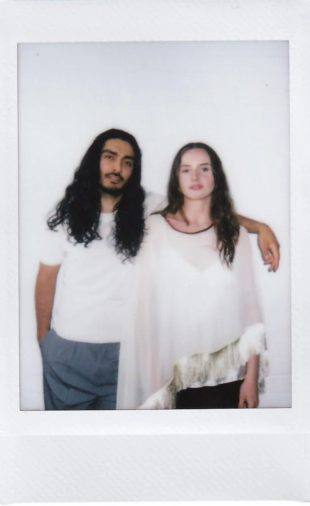 A man and woman standing close together with the man's arm around the woman's shoulder, both looking at the camera, in front of a plain white background.