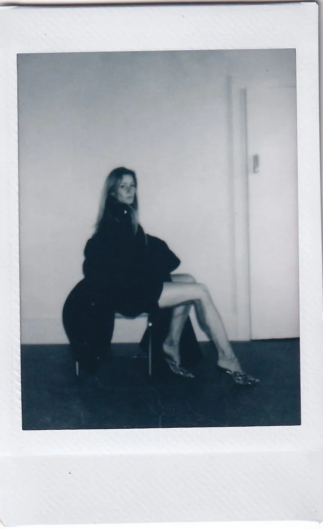A woman sitting on a chair against a plain wall, wearing a dark top and skirt with high heels, in a black-and-white photograph.
