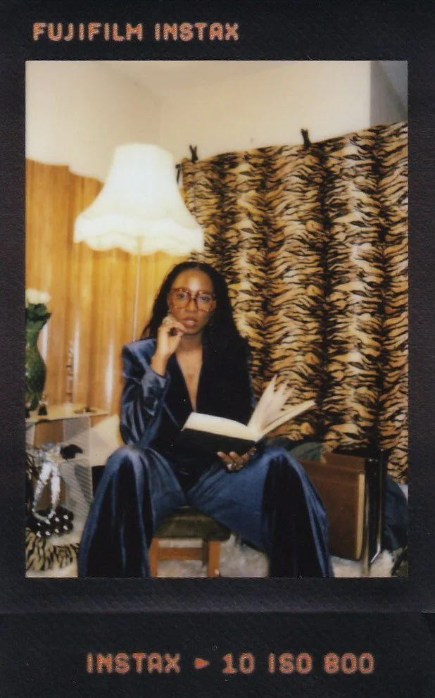 A woman with glasses sitting on a chair, wearing a shiny dark blue suit, holding an open book, in a room with wooden paneling, a zebra-patterned curtain, a tall lamp, and a small table with a flower vase and accessories.