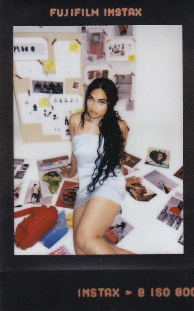 A young woman with long, dark, curly hair sitting on the floor in a white towel or dress, surrounded by photographs and images spread out on the floor and pinned on a wall behind her, with a black border and the words 'Fujifilm Instax' at the top.