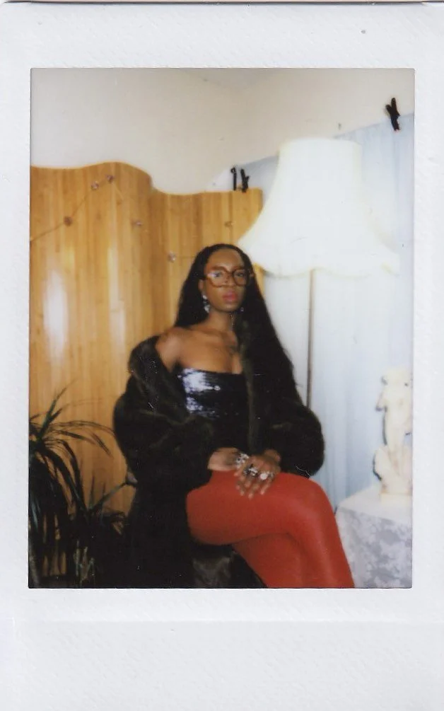 A woman with long black hair and glasses sitting in a room with wooden panel walls, a tall white floor lamp, and a plant in the background. She is wearing a black jacket, a shiny black top, red pants, and has rings on her fingers.