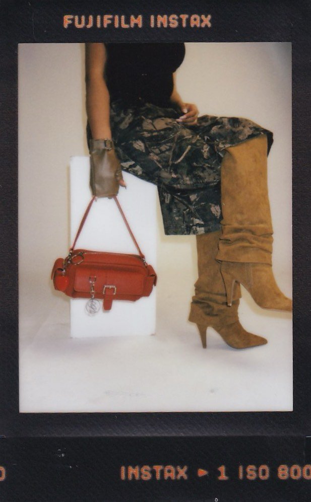 Close-up of a person sitting, wearing a black top, camouflage pants, and tan high-heeled knee-high boots. They are holding a brown purse with a red strap and a hanging keychain, with their right arm resting on their lap.