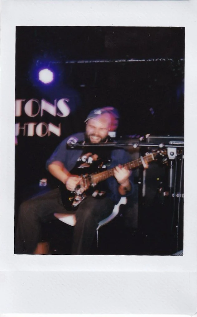 Man playing an electric guitar on stage with a microphone in front, in a dimly lit setting with stage lights and a sign partially visible in the background.