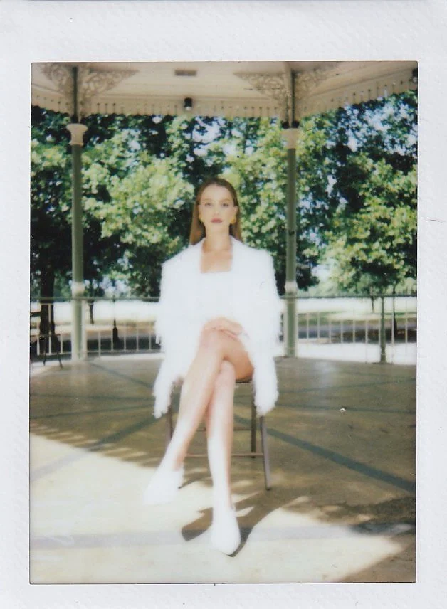 A woman in a white outfit sitting on a chair in a gazebo with trees in the background.