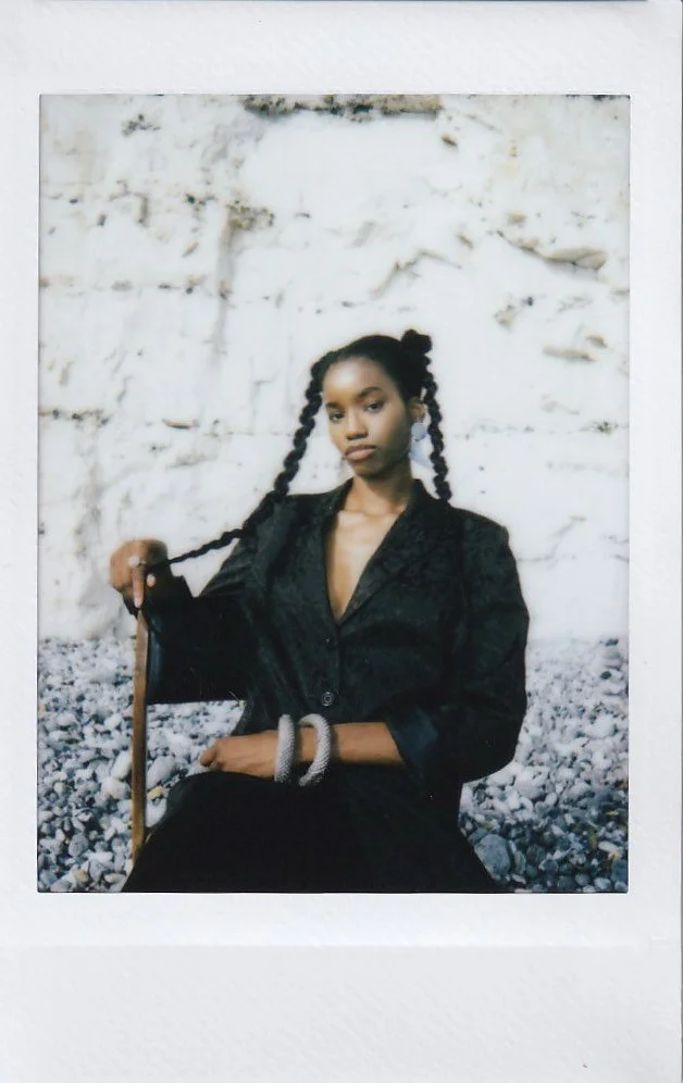 A young woman with dark skin and long braided hair, sitting on a bed of rocks in front of a white brick wall. She is wearing a black blazer and bracelets, looking at the camera.