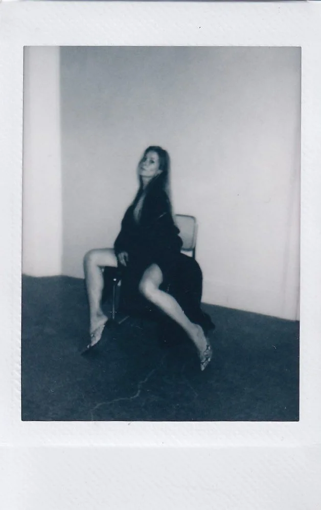 Black-and-white photo of a woman sitting on a chair with one leg crossed over the other, smiling, in a minimal room with plain walls.