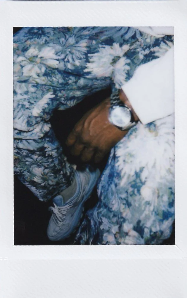 Close up of a man wearing a floral patterned blue trousers, a grey jumper, and a silver watch