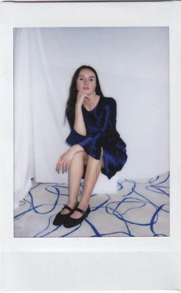 A young woman with long dark hair, wearing a blue satin dress and black shoes, sitting on a white box in a room with white walls and a blue tangled pattern on the floor.