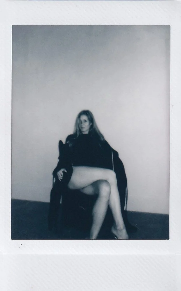 Black-and-white Polaroid photo of a woman sitting on a chair with her legs crossed, wearing a dark sweater and coat, with a plain wall background.